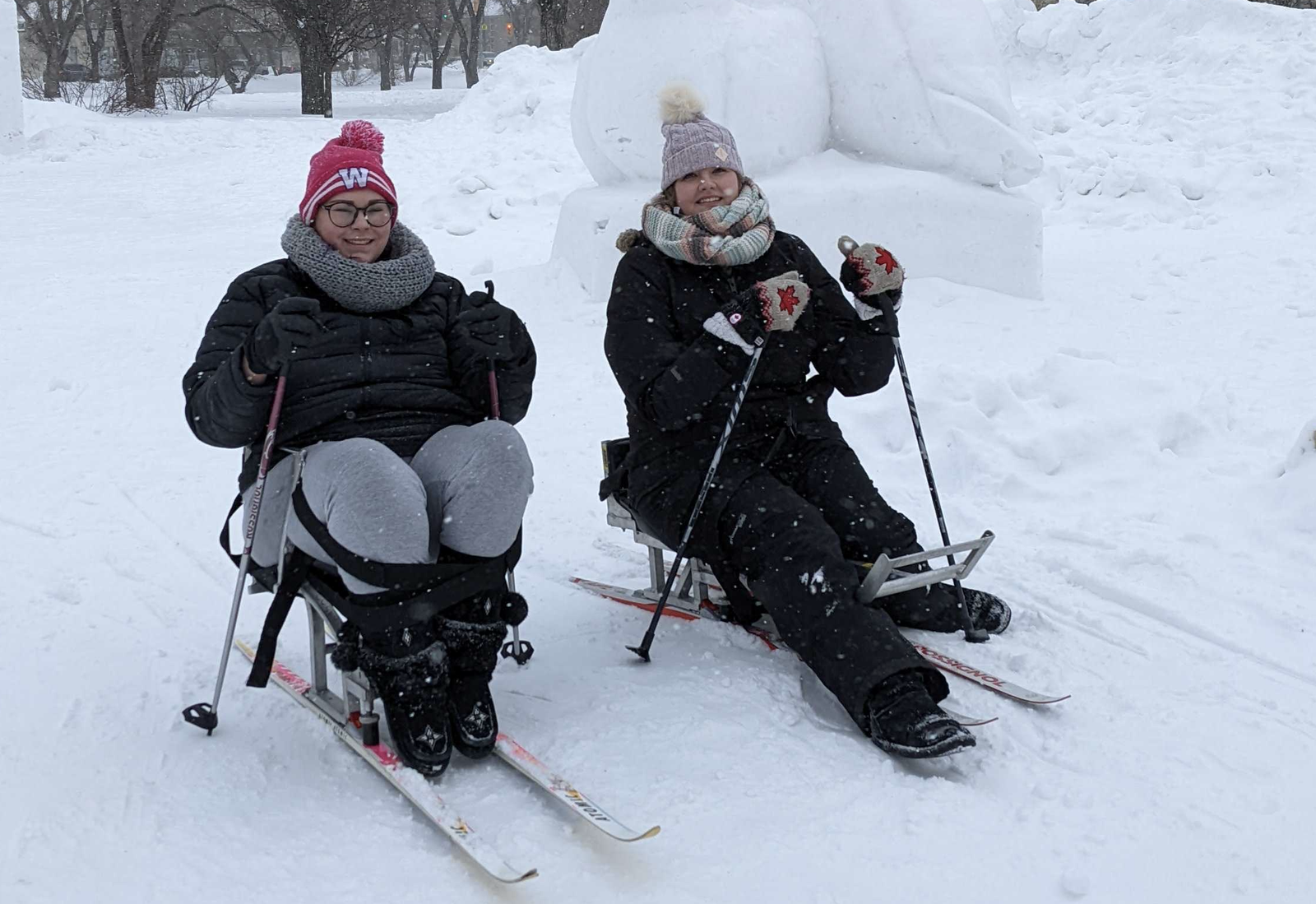 Two women sitting on adaptive sports equipment in a snowy park, dressed warmly with hats and gloves. They are smiling and enjoying the winter weather while surrounded by snow. A large snow sculpture is visible in the background. Winterpeg Adaptive sports.