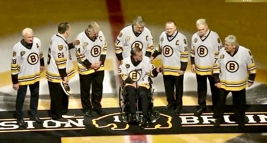 A group of seven men, including a Boston Bruin in a wheelchair, wearing yellow and black jerseys, gathered on an ice rink for a ceremonial event. They are standing on a logo representing the Boston Bruins.