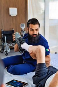 A healthcare professional in scrubs assists a patient lying in bed by adjusting their shoe. A wheelchair is visible in the background, along with a tablet on the bed. This scene depicts rehabilitation to help individuals regain function after SCI.