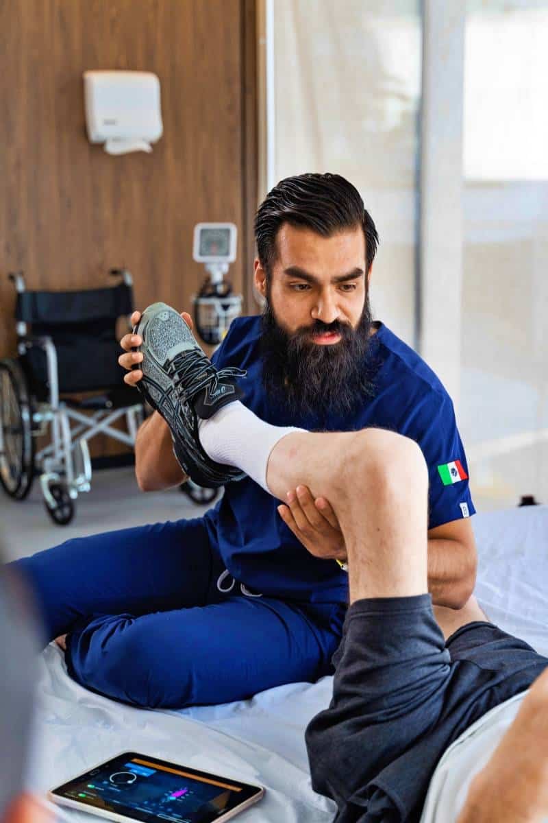 A healthcare professional in scrubs assists a patient lying in bed by adjusting their shoe. A wheelchair is visible in the background, along with a tablet on the bed. This scene depicts rehabilitation to help individuals regain function after SCI.