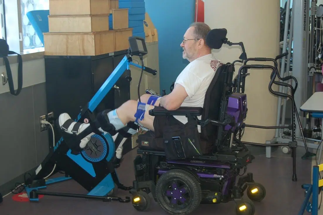 A man using a power wheelchair while engaging with a stationary exercise bike designed for rehabilitation. The setting appears to be a therapy room, with equipment and boxes in the background. This setup showcases State of Art SCI therapy.