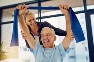 A senior man and a female therapist engaging in a rehabilitation exercise, with the man holding a blue resistance band above his head, smiling. The setting is bright with large windows, indicating a positive environment supporting signs of recovery from SCI.