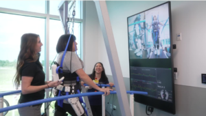 A woman in a robotic exoskeleton supported by harnesses, practicing walking in a rehabilitation facility. Two staff members observe and monitor progress on a screen displaying data related to her movement. The room features large windows providing natural light.