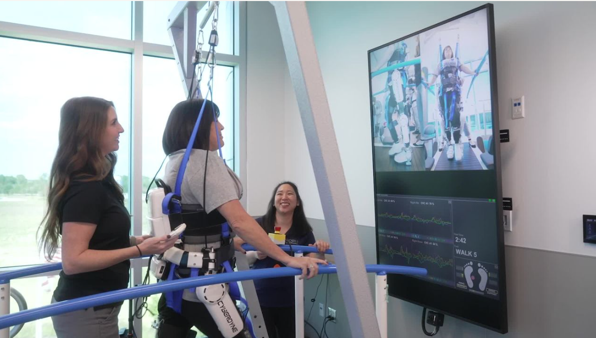 A woman in a robotic exoskeleton supported by harnesses, practicing walking in a rehabilitation facility. Two staff members observe and monitor progress on a screen displaying data related to her movement. The room features large windows providing natural light.