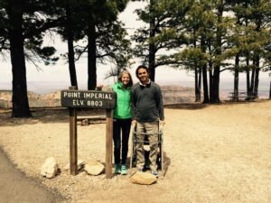 Two people posing by a sign that reads 'Point Imperial ELV 8803' in a forested area, with mountains visible in the background. One person is using a walker, representing inspiring SCI recoveries.