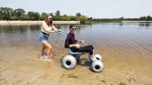 A woman pulling a man in a specialized wheelchair with large wheels through shallow water at a beach, surrounded by trees and a calm landscape, illustrating a Manitoban SCI story.