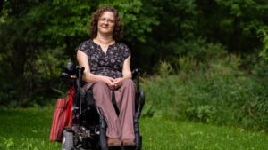 A Métis artist with a disability sitting in a wheelchair on a green lawn, smiling gently. She wears a floral blouse and light pants, with a red and white striped bag beside her. Lush greenery surrounds the scene.