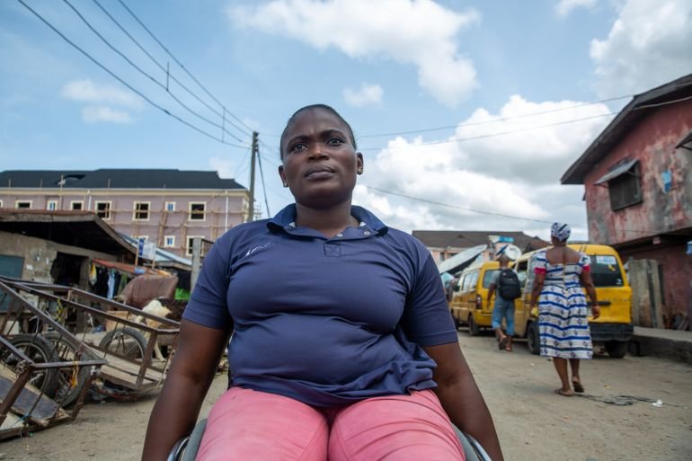 A woman sitting in a wheelchair on a street in Nigeria, dressed in a blue shirt and pink pants, with buildings and pedestrians in the background, highlighting Nigeria life with disability.