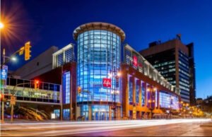 The Canada Life Centre, a modern building with large glass windows, is illuminated at night. Street lights and traffic signals are visible, along with nearby city buildings.