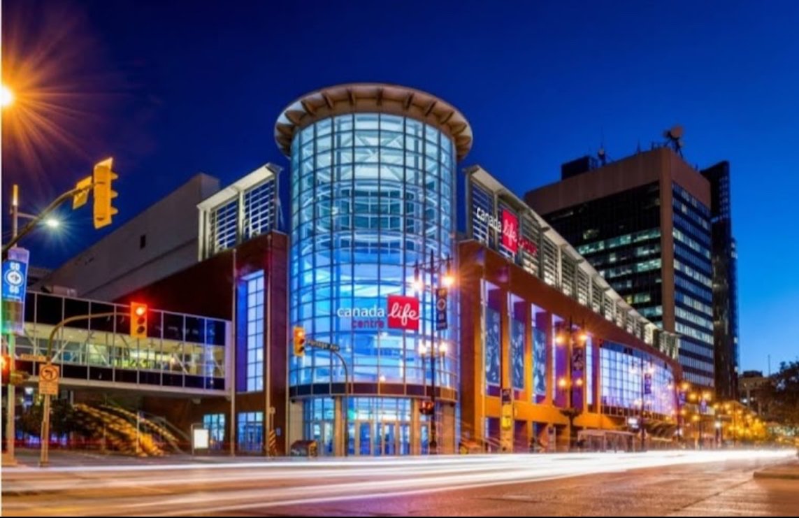 The Canada Life Centre, a modern building with large glass windows, is illuminated at night. Street lights and traffic signals are visible, along with nearby city buildings.