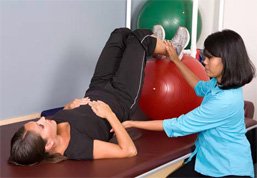 A woman lying on an exercise table with her legs elevated on a ball, while a therapist assists her by holding her foot. Various exercise balls are visible in the background.