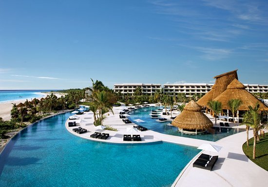 A luxurious resort featuring a large swimming pool with lounge chairs, palm trees, and thatched-roof structures. The ocean is visible in the background under a clear blue sky.
