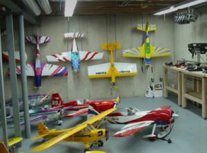 An indoor workshop featuring various model airplanes displayed on a concrete wall and placed on the floor. The colorful designs include an assortment of flying models, showcasing a range of styles and sizes. A workbench is visible in the background, typical for a Model Airplane Club setup.