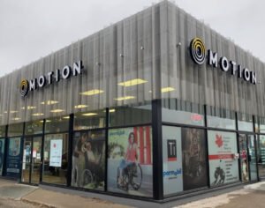 Exterior view of a Motion Canadian Health Care Products store with large windows displaying images of people using mobility aids. The store features a modern design with the "MOTION" logo prominently displayed above the entrance.