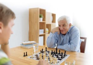 An older man and a young man playing chess at a wooden table. The older man is smiling and resting his chin on his hand, while the young man looks focused on the game. A glass of water and books are visible in the background.