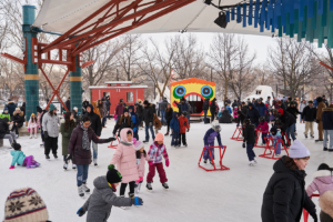 A busy outdoor ice skating rink filled with children and adults in winter clothing. Some skaters use assistance frames while others glide on the ice. Colorful structures are visible in the background. Families enjoy leisure time together.