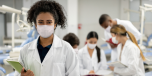 A group of dental students wearing lab coats and masks, with one student smiling in the foreground holding a notebook. Others are engaged in discussion and note-taking at a dental clinic.