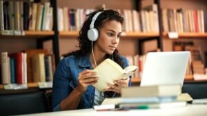 A woman wearing headphones is reading a book while looking at a laptop in a library filled with bookshelves. She has curly hair and is focused on her study.