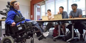 A young man in a power wheelchair speaks in a classroom, while three students sit around a table with laptops, listening attentively. Windows in the background allow natural light into the space.