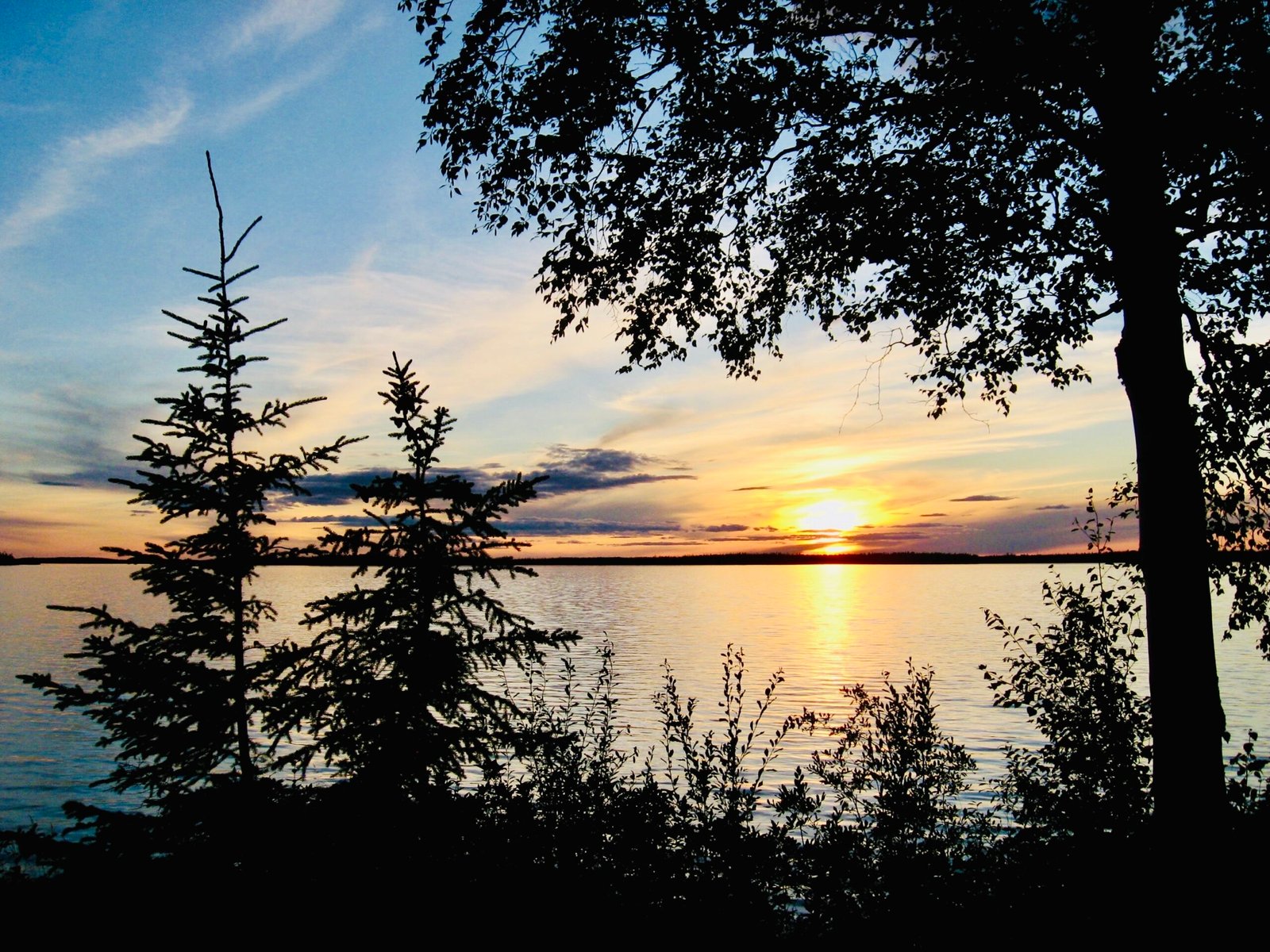 A serene lakeside view at sunset, with silhouettes of trees against a colorful sky. The sun is setting on the horizon, casting warm hues over the water. Experiencing Momenta Outdoors.