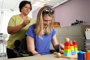 A woman in a wheelchair assembling colorful building blocks on a table, while another woman, standing behind her, offers assistance. The setting is bright and modern, with cabinets in the background.