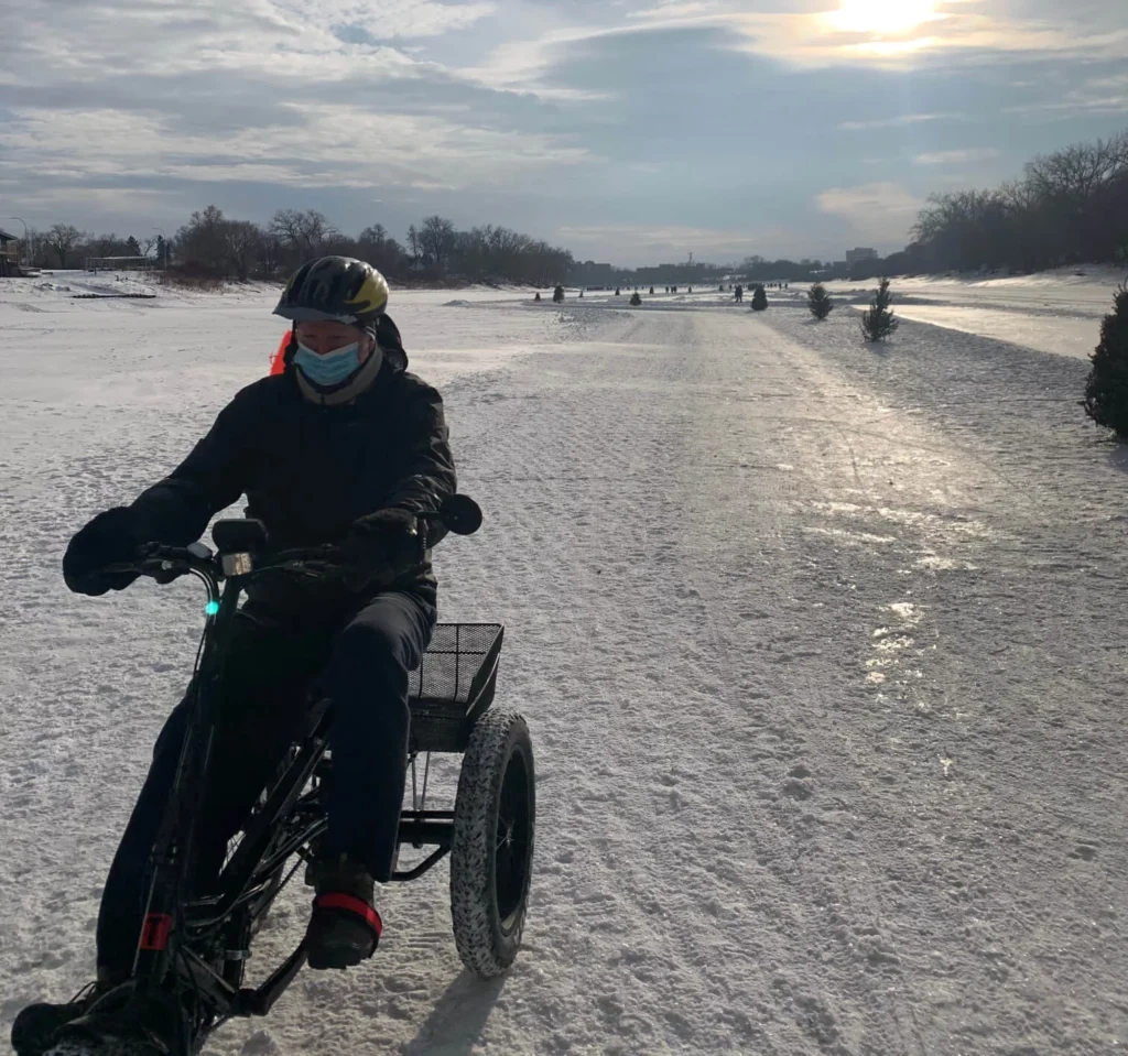 A person wearing a black jacket and mask riding a three-wheeled bike on a snowy path with trees in the background. The sun is low in the sky, casting shadows on the snow-covered ground.