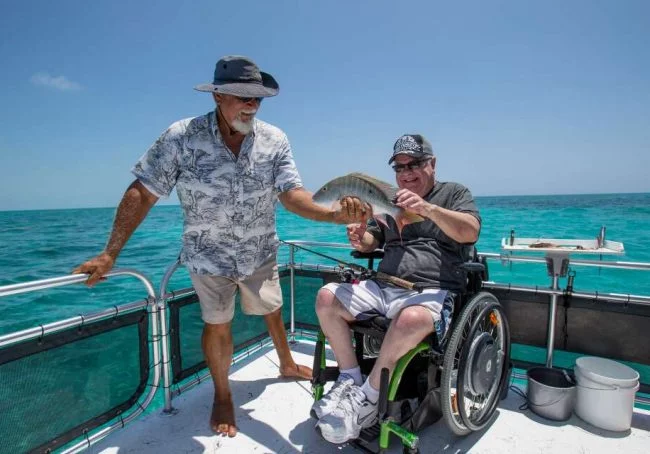A man in a wheelchair smiling and holding a fish, while another man in a hat stands beside him on a boat. The background shows clear blue water and a sunny sky.