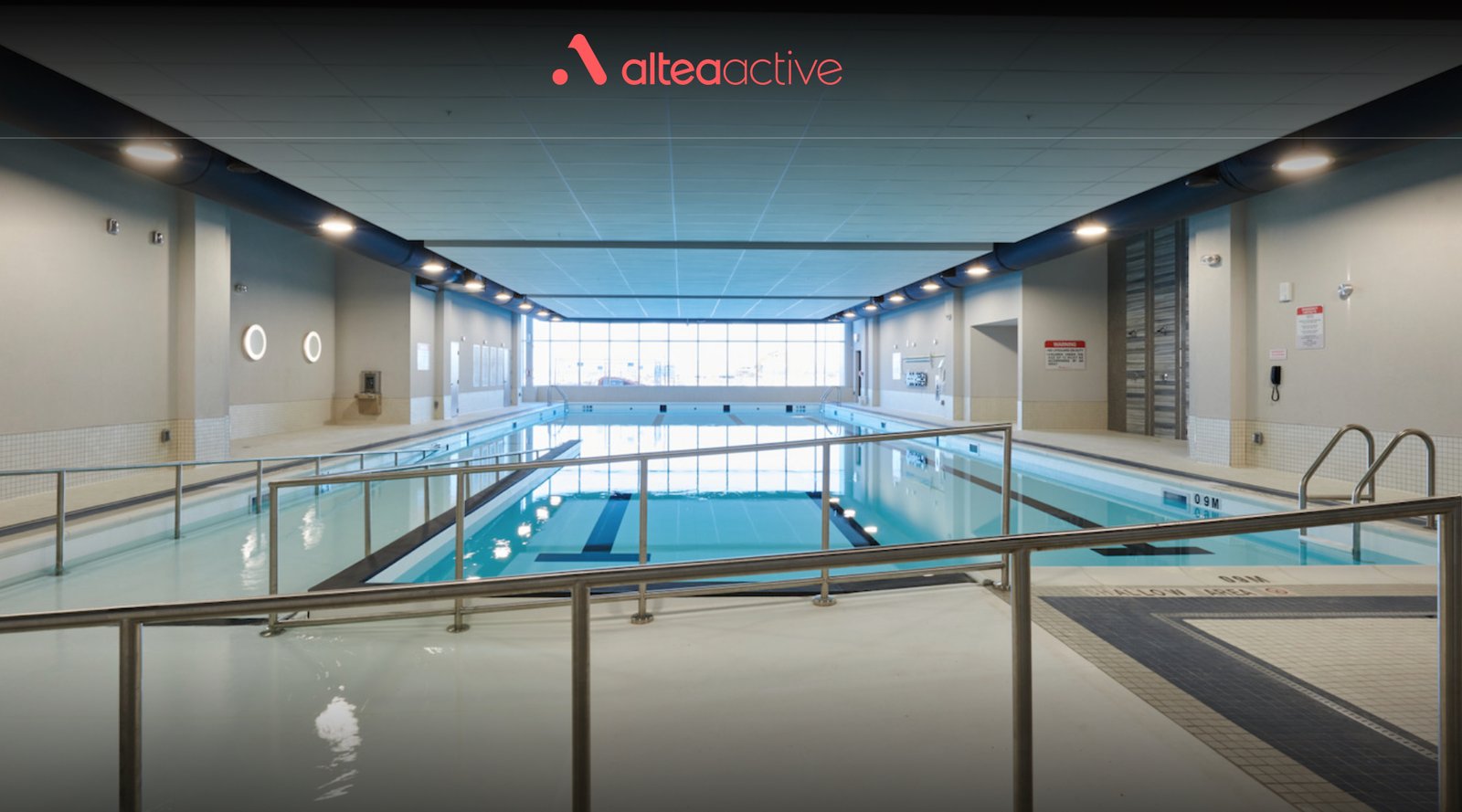 An indoor swimming pool at Altea Active Gym featuring clear blue water, a spacious area, and large windows allowing natural light to enter. A railing runs along the poolside.