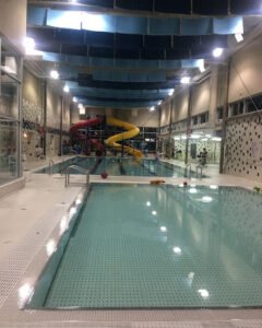 An indoor swimming pool area featuring clear water, a water slide with yellow and red spirals, and a chairlift for accessibility. The environment is well-lit with high ceilings and blue fabric panels above.