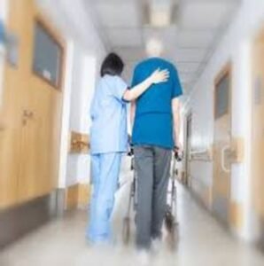 A healthcare worker assisting an elderly patient using a walker in a hospital corridor. The worker has one hand on the patient's back for support, illustrating care and support in Primary Care Staffing.