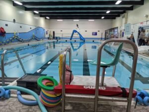 Indoor swimming pool at Transcona Kinsman Pool, featuring clear blue water, lane markings, and poolside equipment. A lifeguard chair with safety gear is positioned nearby, and several people are in the background.