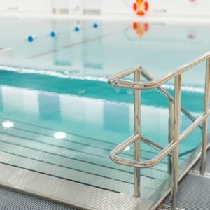 A close-up of a stainless steel pool railing beside a clear blue pool at Aqua Essence Swim Academy, with floating buoy markers visible in the water.