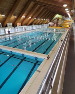 An indoor swimming pool at the Elmwood Kildonan Pool, featuring clear water, lane markings, and a lifeguard on duty. Natural wood beams adorn the ceiling, and a waterslide is visible in the background.