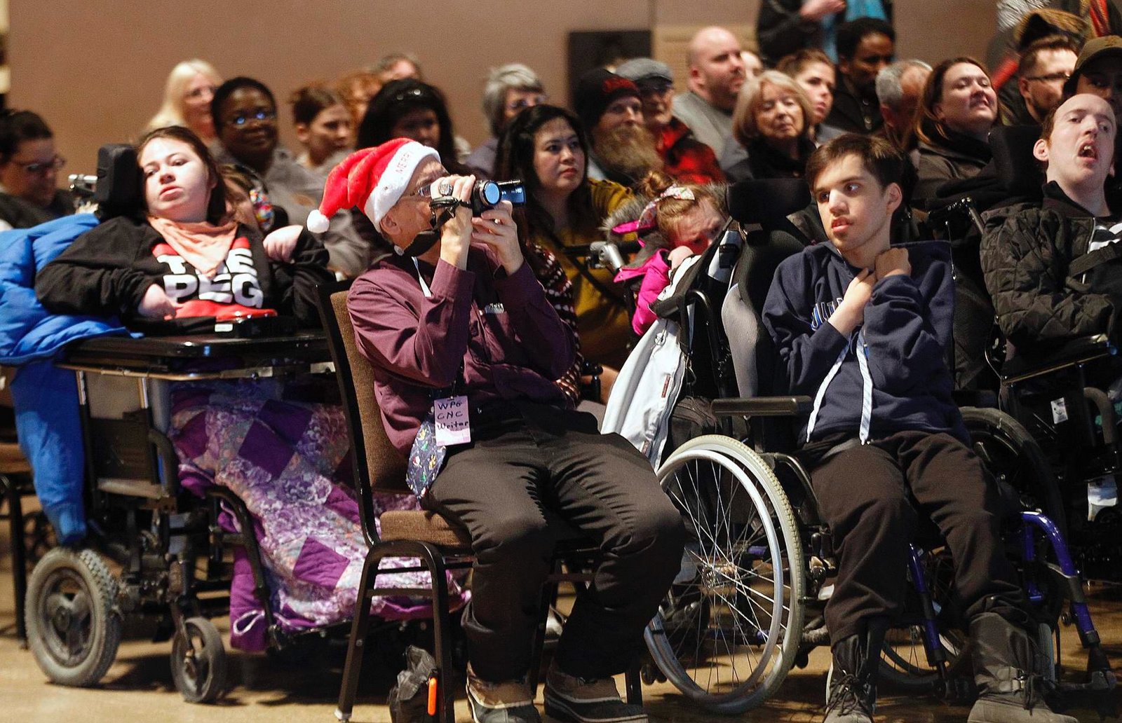 A diverse group of people, including individuals in wheelchairs, seated in an audience at an event. One person wears a Santa hat and holds a camera, while others are engaged in the presentation. The setting suggests a communal gathering, promoting inclusivity.