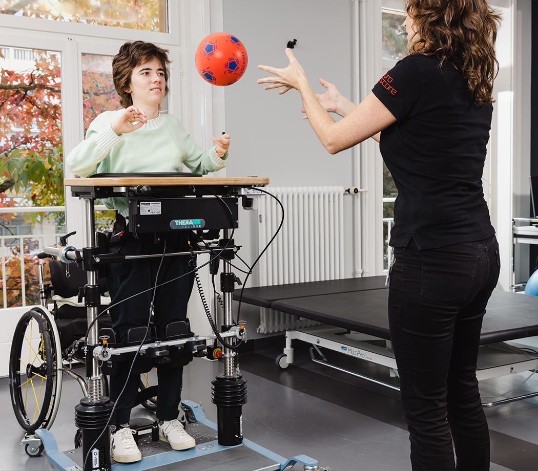 A young woman using a standing wheelchair interacts with a therapist who is tossing a red ball to her in a therapy room. The setting features large windows with foliage visible outside.