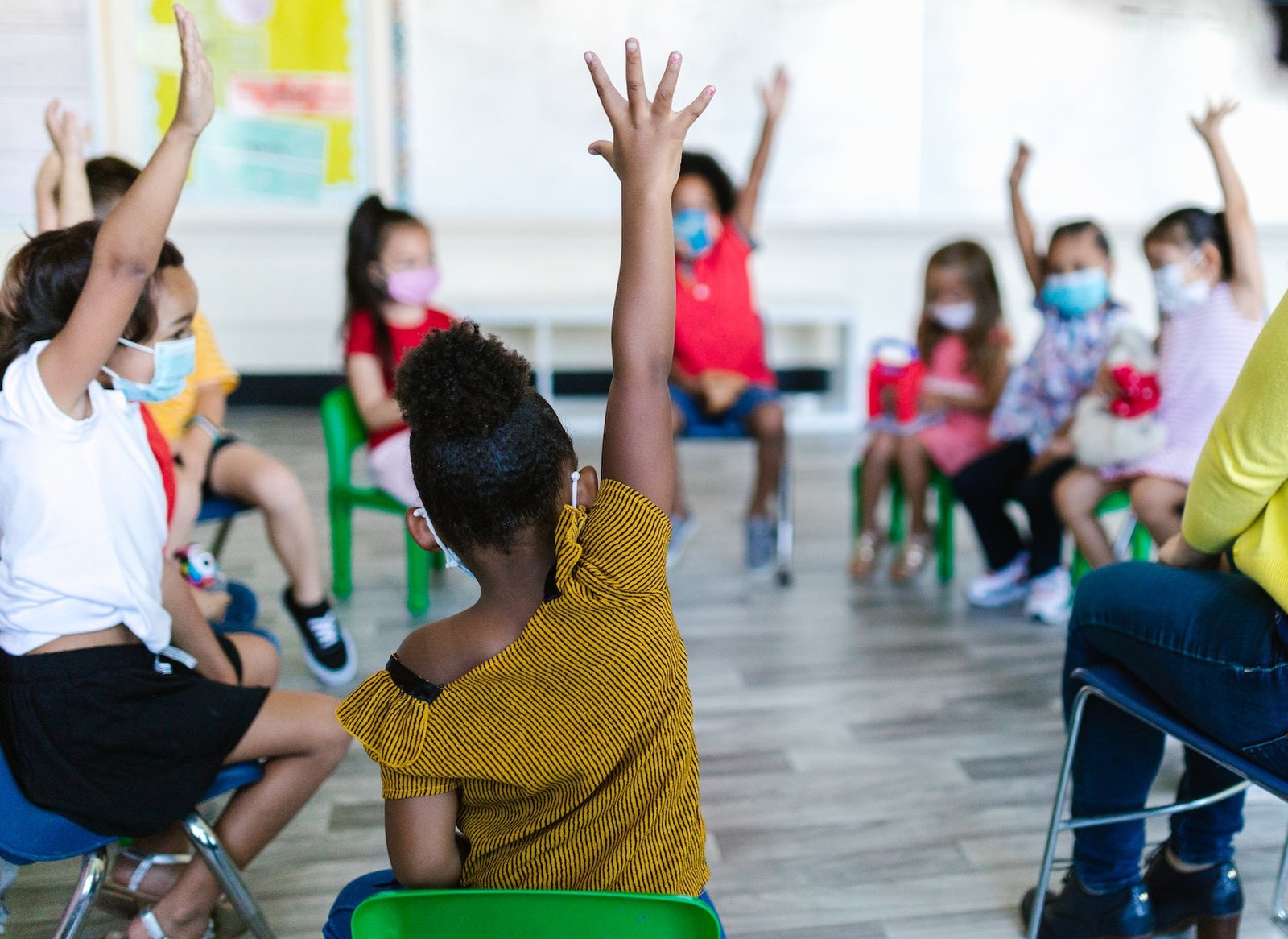 A group of children wearing masks in a classroom, engaged in a discussion with hands raised. The focus is on a girl with curly hair and a yellow striped shirt, sitting on a green chair, participating actively. Think First: Students Accident Prevention.
