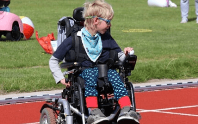 A child with blond hair wearing sunglasses and a light blue scarf is seated in a wheelchair on a running track. The environment includes green grass and various colorful items in the background, suggesting a playful outdoor setting for Gympanzees Kids Exercises & Games.