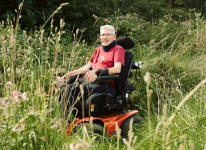 A person in a red shirt sitting in a motorized wheelchair among tall grass and wildflowers in a natural setting. The background features greenery and shrubs, illustrating a serene outdoor environment. The image relates to "30 Dying People Share What’s Important."