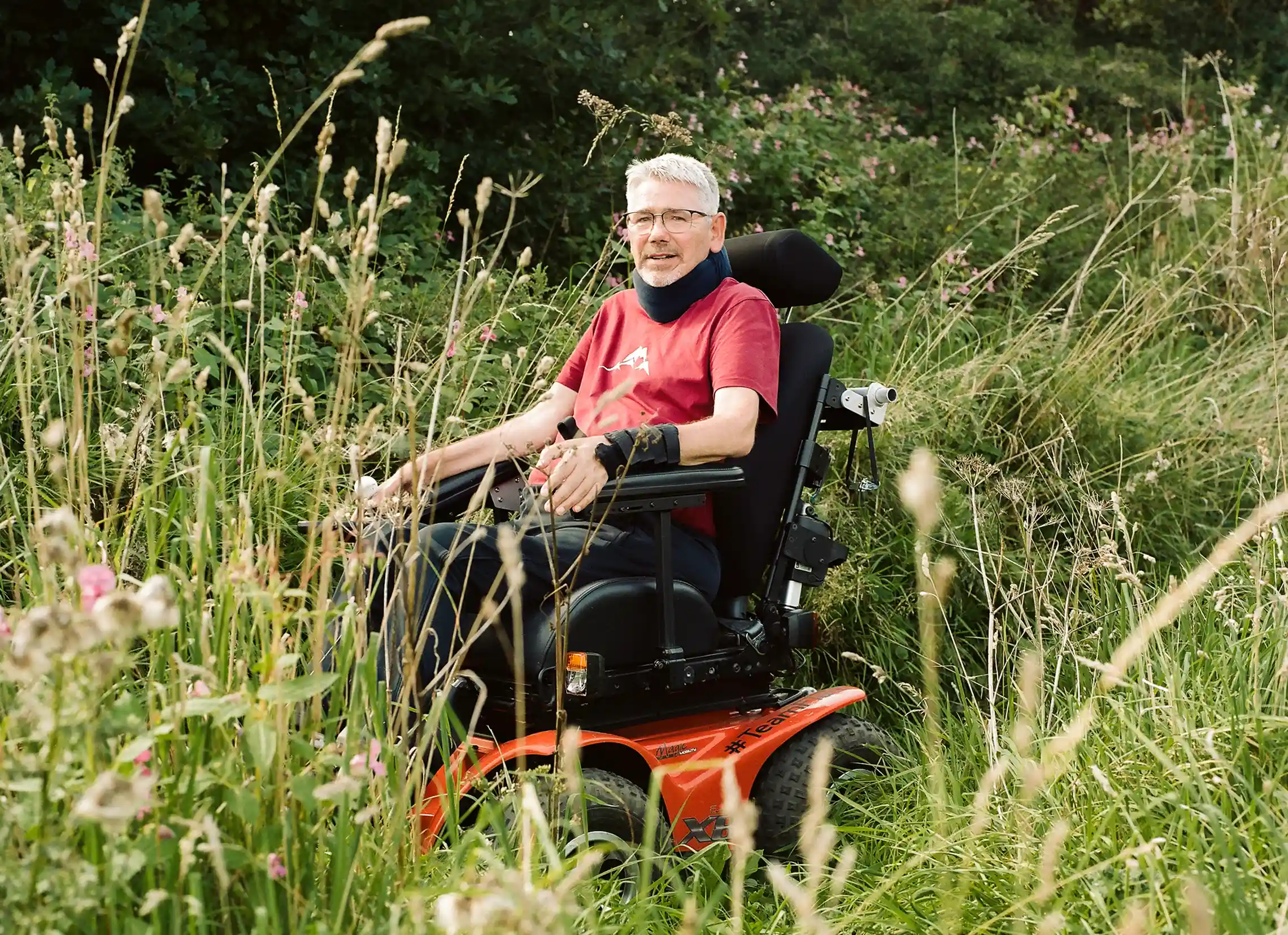 A person in a red shirt sitting in a motorized wheelchair among tall grass and wildflowers in a natural setting. The background features greenery and shrubs, illustrating a serene outdoor environment. The image relates to "30 Dying People Share What’s Important."