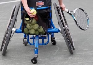 A person in a wheelchair holding a tennis ball while other tennis balls are in a mesh bag attached to the back of the chair, with a tennis racket in the other hand, on a court.