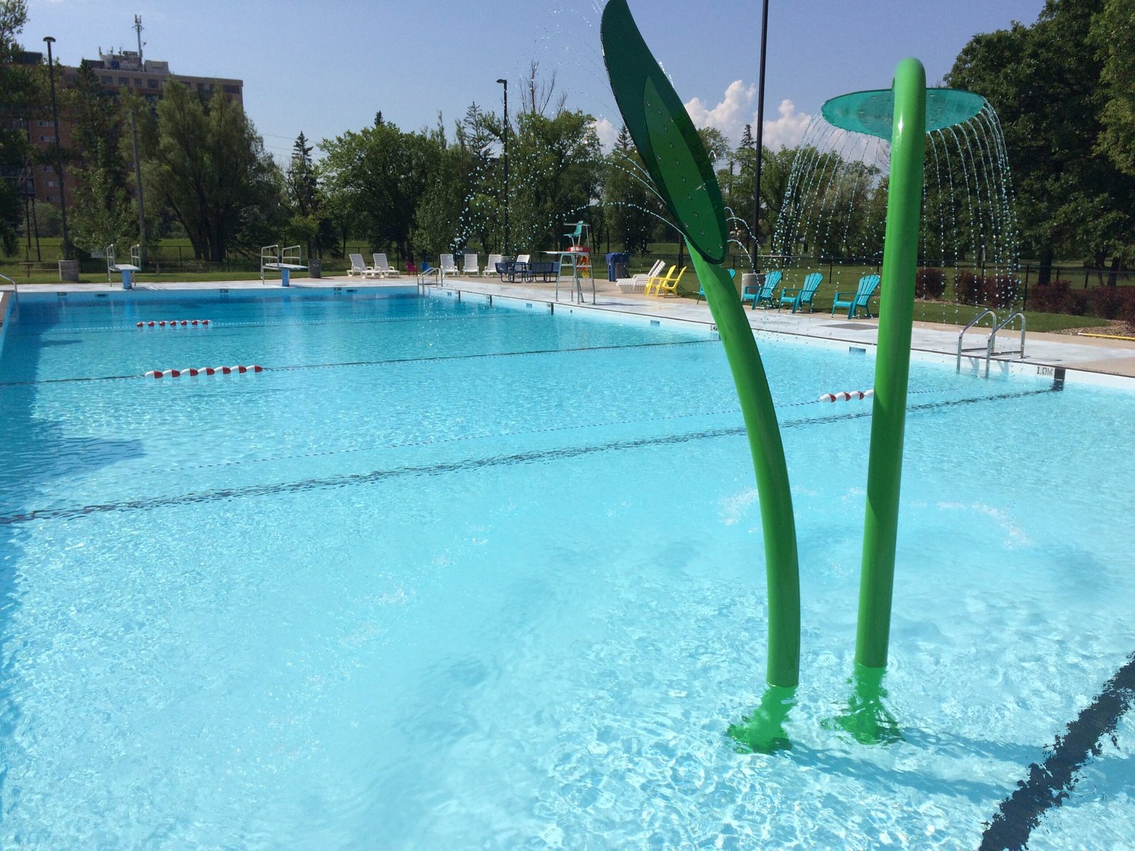 A bright outdoor swimming pool featuring beach entry, with green water slide structures resembling plants. Lounge chairs are set up around the pool area and surrounded by trees, under a clear blue sky.