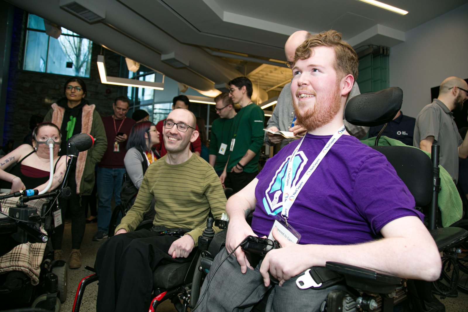 A group of individuals at an event, with two men in wheelchairs smiling and engaging with others. They are surrounded by attendees in a modern space, showcasing collaboration and community at 'Makers Making Change - Assistive Technology'.