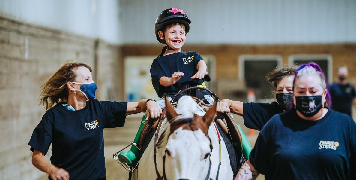 A child wearing a helmet smiles while riding a horse, assisted by three adults wearing masks. The adults are supporting the child on both sides in an indoor arena. The child and adults wear matching shirts featuring "Prairie Strides Adapted Horseback Riding."