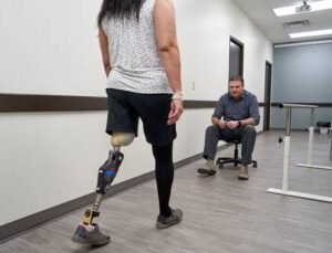 A woman with a prosthetic leg walking in a rehabilitation room while a man observes from a chair. The setting includes a plain floor and walls, with equipment for physical therapy visible in the background. Manitoba Orthotics & Prosthetics is relevant to the context.