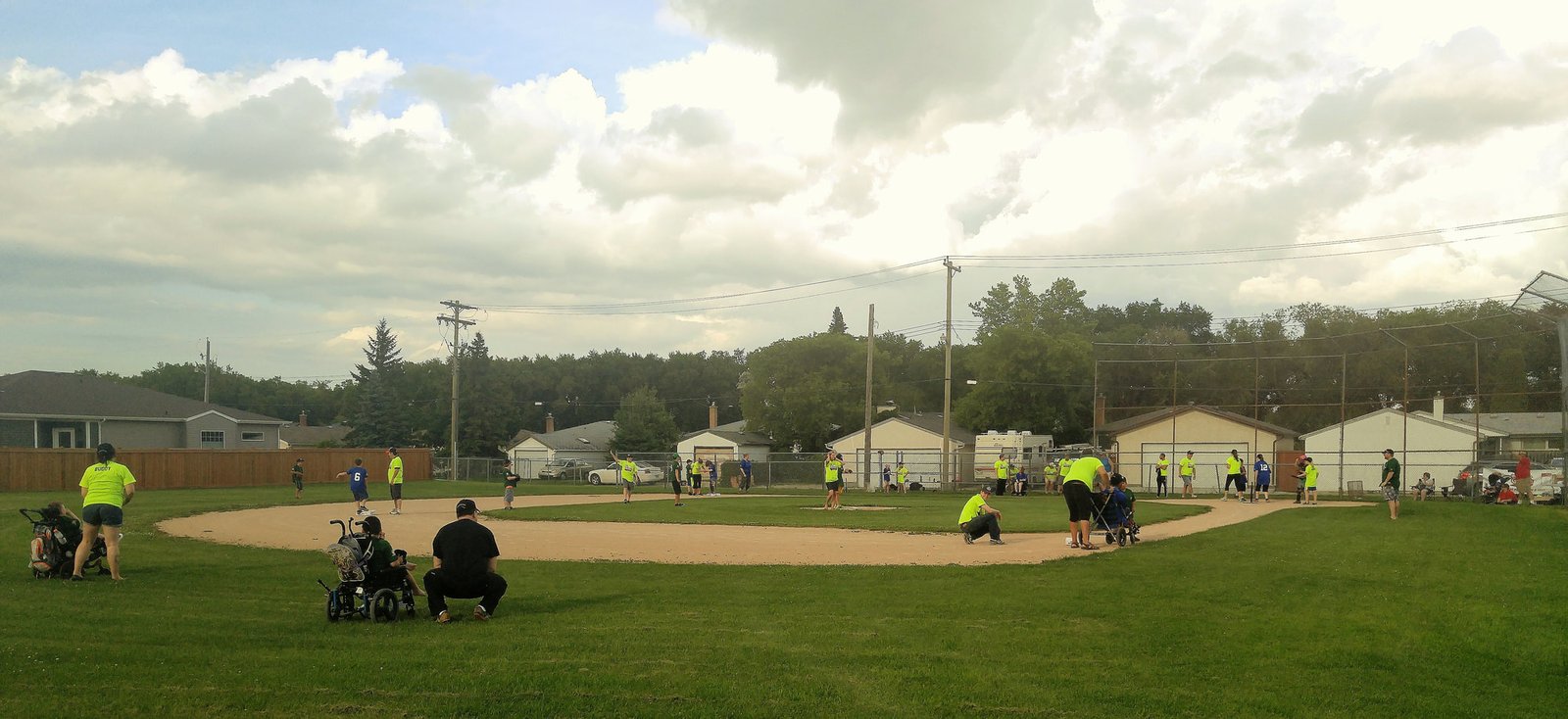 A community baseball field with players in yellow shirts engaged in a game, and spectators in the foreground. The background features houses and trees, under a cloudy sky. This scene is related to Challenger Baseball Manitoba.