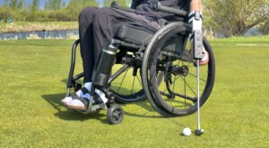 A person in a wheelchair positioned on a golf course, holding a golf club while preparing to putt. The individual is wearing casual black attire and adaptive features are visible on the wheelchair, highlighting C5 Mobility Adaptive Products.