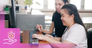 Two women seated at a desk, one assisting the other at a computer. The woman on the right is using the keyboard while the woman on the left smiles. The image includes a graphic of a hand and a star on the left. This visual represents guidance and support related to how to apply for disability supports in Manitoba.