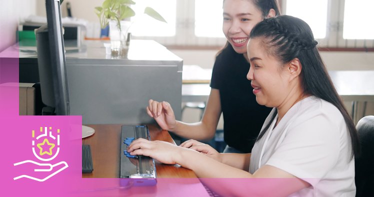 Two women seated at a desk, one assisting the other at a computer. The woman on the right is using the keyboard while the woman on the left smiles. The image includes a graphic of a hand and a star on the left. This visual represents guidance and support related to how to apply for disability supports in Manitoba.