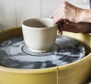 A person's hand shaping a beige pottery bowl on a spinning pottery wheel. The background features a blurred workspace with tiles.
