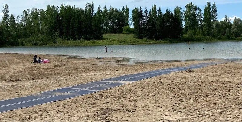 A sandy beach area with a blue accessibility mat leading to a calm body of water. In the background, people are enjoying the water, and trees line the shore, providing a natural setting. The image highlights accessibility mats on Manitoba beaches.